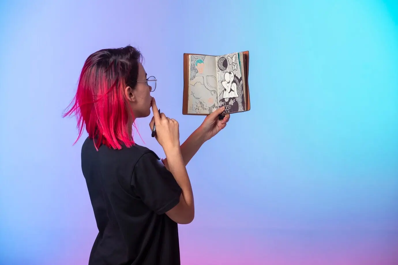 Юная девушка с розовыми волосами держит скетчбук и смотрит на что-то.